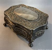 Photo of vintage metal rectangular jewelry box, embossed decorative patterns on lid and sides, on claw feet, with monogram initials on top.