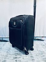 Black soft fabric suitcase standing upright with extended telescopic handle and four spinner wheels visible. Front zippered compartments and brand logo patch visible.