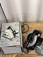 Front view of the circular saw next to its original box, power cord visible, extra blades and wrench present on the table.