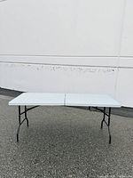 Full view of table unfolded outdoors on pavement, showing white plastic top and black legs.