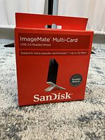 Front view of the red retail box of SanDisk ImageMate Multi-Card USB 2.0 Reader/Writer. Shows product image, SanDisk branding, and key features including transfer button and 30 MB/s speed.