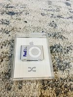 Front view of the sealed silver Apple iPod Shuffle with white circular control pad inside clear plastic packaging, resting on carpet.