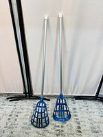 Two complete lacrosse sticks side by side showing blue plastic heads with grid design and white shafts, standing vertically against a white backdrop.