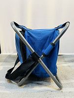 Blue camping stool shown folded with black adjustable strap and silver metal tubular frame visible, fabric is intact.