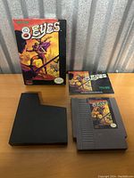 Photo showing the 8 Eyes complete game lot with box, cartridge, plastic case and manual displayed on table.