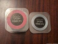 Two rolls of satin ribbon, red and silver, in square plastic packaging labeled 'waste not paper SATIN RIBBON' on wood surface.