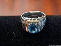 Close-up photo of men's sterling silver ring with cushion cut blue topaz gemstone in center, resting on a black surface.