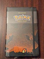 Front cover of the black Moleskine journal showing the Pokemon logo and a stylized orange Charmander with flame tail in dot pattern.