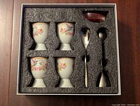 Four porcelain soft boiled egg cups with floral and bird design, two double-ended metal spoons, and a small ceramic dish in foam packaging inside decorative box