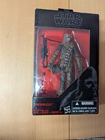 Front view of unopened Star Wars The Black Series Chewbacca figure with bowcaster accessory visible inside clear plastic casing, black and red themed box.
