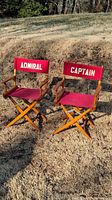Two wooden folding director chairs side by side on grass. One labeled 'ADMIRAL' and the other 'CAPTAIN' on red fabric backrests.