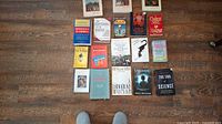 Overview of 16 softcover and paperback books arranged on a wooden floor, showing front covers and titles of most books.