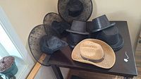 Photo showing 4 black witch hats, 10 black top hats, and 1 tan straw cowboy hat on a table.