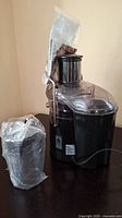 Black electric juicer with transparent lid and power cord, wrapped in plastic.