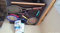 Full view of all sports rackets and balls placed on a black surface and cardboard box.