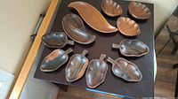 Overview of all wooden serving bowls and platters on a dark table. Various curved shapes, some with handles resembling fruits or pods, others without handles.