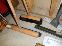 Two push brooms with wooden handles and natural bristles resting on floor, rubber squeegees and part of a mop visible nearby.