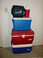 Multiple coolers stacked including two Igloo coolers (red and blue) and a blue Coleman cooler with white lid along with the Playmate thermal jug (red and white) and black AmeriServet insulated bag on top.