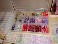 Close-up photo of multi-colored beads organized in clear compartmentalized plastic boxes showing variety of shapes and colors including red, purple, pink, green, and pearl beads.