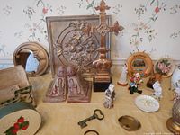 Photo showing fruit motif wall plaque, gold acorn bookends, round moon mirror, wood cross statue, red wood photo album, ceramic figurines, and assorted small decorative items on a table against floral wallpaper.