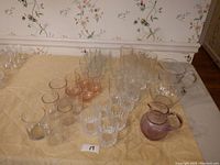 Photo showing the full lot of drinking glasses arranged in rows on a cloth surface, with crystal and colored glasses visible alongside two different pitchers.