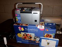 Epson PictureMate printer displayed on top of the original box with user manual and extra ink cartridge box next to it. Shows all included items in the lot.