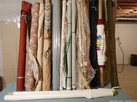Side view of 13 rolled fabrics varying in colors and patterns including florals, damasks, solids, and geometric patterns wrapped on cardboard tubes.