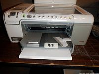 Front angled view showing paper tray extended, control buttons, and USB cable inside output area with lot number tag