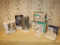 Front view of the three humidifiers on a table, showing the Duracraft humidifier with box and replacement filters, the silver Air Innovations humidifier, and the KAZ Reli-On humidifier stacked on its box.
