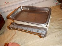Vintage metal household scale with a rectangular metal tray and slider weights on display, showing the scale platform and rod mechanism beneath.