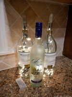 Three wine bottles on a kitchen counter with lights inside; two clear Woodbridge bottles wrapped with plug-in string lights and one frosted Vineyard Estates bottle with battery light and blue screw cap. Battery pack visible.