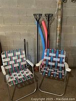 Photo showing 2 vintage webbed lawn chairs, 2 wrought iron planter stands, 2 pool noodles, and a rolled bamboo shade leaning against a concrete block wall.