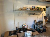 Photo of cardboard boxes containing various black plastic irrigation parts and planter components, some stacked plastic pots and containers, and miscellaneous gardening materials on the floor beneath a wall-mounted metal shelf.