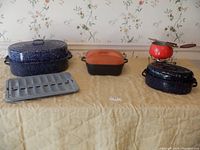 Overview of 5 specialty cooking pans on table, showing large speckled roaster, orange fondue pot with forks, small black/white roaster, metal drainer.