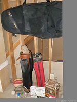 Two golf bags, one black with a cover and one red with a plush head cover, accompanied by a deflated basketball and other sports balls on the floor, tennis racket leaning against golf bags, grill basket and other grilling items on floor