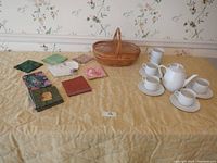 Photo showing all items in set: Wunsiedel Bavaria porcelain tea set including teapot, 7 cups (5 with saucers, 2 extra), decorative paper napkins, and one-handled wicker basket on yellow quilted surface.