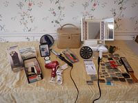 Photo shows full spread of vintage grooming lot on a bed with beige cover, floral wallpaper background. Items visible include train case, lighted mirror, multiple hair dryers, boxed shaver, round and rectangular mirrors, curling iron, combs, and barber tools.