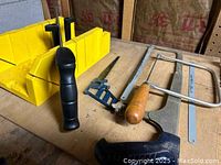 Yellow plastic miter box with black-handled hacksaw and small metal handsaw with wooden handle shown on work surface