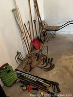 Wide view showing the assortment of yard tools leaning against a wall, with the red plastic shovel, wooden handles, sickles, and green garden hose clearly visible.