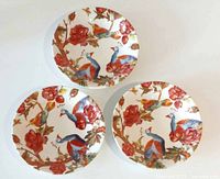 Top-down view of three ceramic bowls featuring colorful peacock and floral designs in blue, orange, and red tones on white background.
