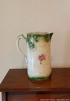Side view of antique ceramic pitcher with floral decoration and green embossed details, capturing the pink single flower motif and crackled glaze.