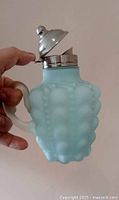 Photo showing the light blue satin glass syrup pitcher with embossed bubble pattern and hinged metal lid partially open held by a hand to show size and detail.