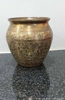 Front view of antique brass and copper water pot vase showing detailed floral engravings and geometric border pattern near rim on dark surface.