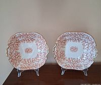 Front view of two Minton cake plates with red coral seaweed patterns, displaying shape, handles, and gold trim.