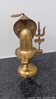 Photo shows full view of brass Shiva Lingam with cobra hood and trident on flat surface indoors
