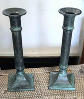 Pair of cast bronze neoclassical candle holders with square base and verdigris patina on a patterned surface.
