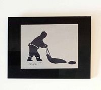 Frontal photo of the Inuit block print art showing a figure pulling a sled on white paper with black plastic backing.