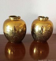 Pair of rounded solid brass vases with engraved Japanese landscape scenes, displayed side-by-side on a wooden surface.