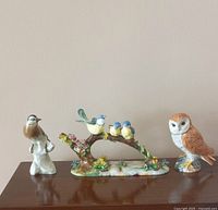 Three porcelain bird figurines arranged on a wooden surface against a plain wall: a small brown bird on a white base, a three birds on a branch larger colorful bone china piece, and a barn owl figurine.