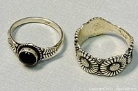 Two vintage sterling silver rings on white textured surface, showing overall styling with one ring featuring a black stone and feather-style engraved band, the other a wide band with floral sunflower pattern.